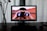 A computer monitor displays an image of a person looking through binoculars with Facebook logos on the lenses. The setup includes a desktop and a glass vase with long, dark plant stems on a wooden table, with curtains in the background.