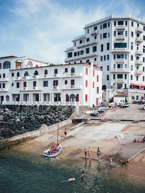 A coastal scene features several multi-story white buildings with balconies and red accents. People are gathered by the water, some swimming or playing near a small boat on a concrete ramp. The setting looks like a seaside town and conveys a leisurely, vacation-like atmosphere.