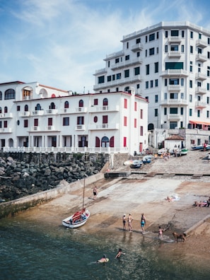 A coastal scene features several multi-story white buildings with balconies and red accents. People are gathered by the water, some swimming or playing near a small boat on a concrete ramp. The setting looks like a seaside town and conveys a leisurely, vacation-like atmosphere.