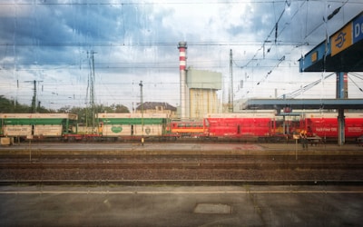 Several trains are stationary on railway tracks, with overhead electric wires stretching across the top of the image. The trains are primarily red and green and are situated near a platform with a sign partially visible on the right. There is a large industrial structure with a tall, red and white striped chimney in the background, under a partly cloudy sky.