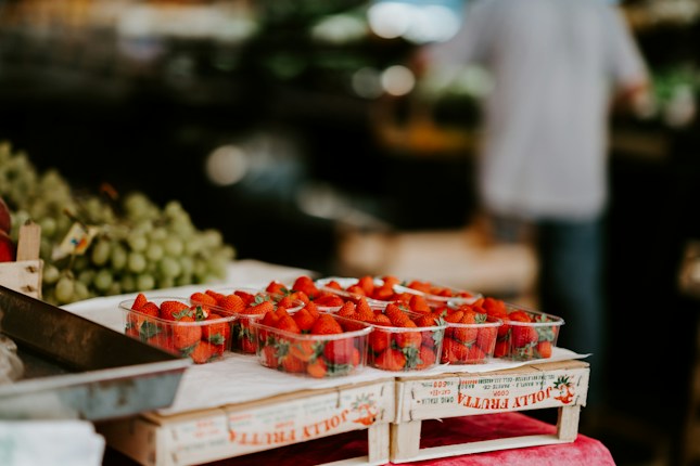 A display of fresh strawberries arranged neatly in transparent containers placed on a wooden crate at a market. In the background, there are bunches of green grapes and blurred figures, suggesting an active market environment.