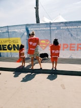 Three people are standing near a chain-link fence with promotional banners. Two children are wearing red t-shirts, and an adult is between them. There is a large logo resembling Superman's emblem in the center of the banner.