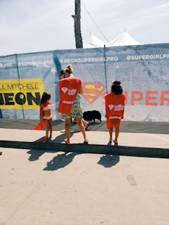 Three people are standing near a chain-link fence with promotional banners. Two children are wearing red t-shirts, and an adult is between them. There is a large logo resembling Superman's emblem in the center of the banner.