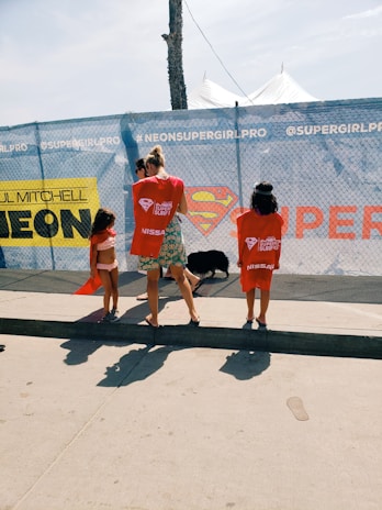 Three people are standing near a chain-link fence with promotional banners. Two children are wearing red t-shirts, and an adult is between them. There is a large logo resembling Superman's emblem in the center of the banner.
