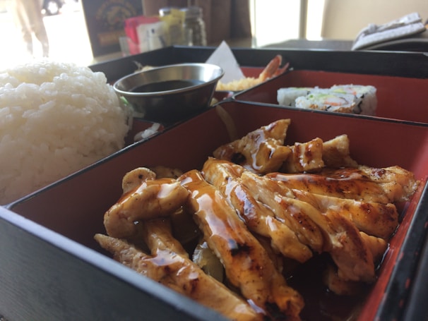 The image features a bento box with grilled chicken drizzled with sauce, a serving of white rice, sushi rolls, and a small bowl of dipping sauce. The bento box is open, revealing the food arranged neatly inside.