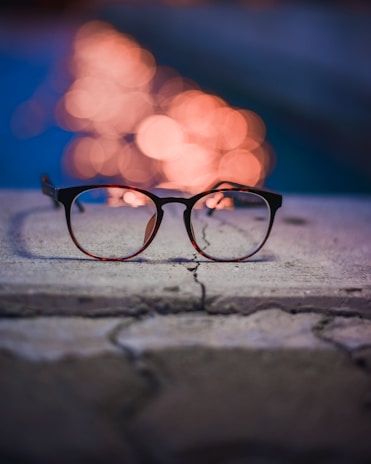 A pair of eyeglasses rests on a textured stone surface, with a backdrop of blurred, luminous orange lights against a deep blue background, creating a bokeh effect.