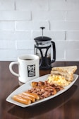 A hearty breakfast consisting of sausages, scrambled eggs, bacon, corned beef, and toast, served on a rectangular white plate. Accompanying the meal is a mug of coffee and a French press containing more coffee. The setup is placed on a dark wooden table against a white brick wall background.