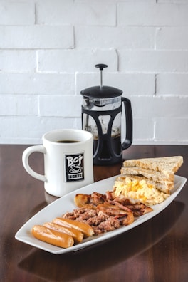 A hearty breakfast consisting of sausages, scrambled eggs, bacon, corned beef, and toast, served on a rectangular white plate. Accompanying the meal is a mug of coffee and a French press containing more coffee. The setup is placed on a dark wooden table against a white brick wall background.