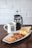 A hearty breakfast consisting of sausages, scrambled eggs, bacon, corned beef, and toast, served on a rectangular white plate. Accompanying the meal is a mug of coffee and a French press containing more coffee. The setup is placed on a dark wooden table against a white brick wall background.