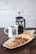 A hearty breakfast consisting of sausages, scrambled eggs, bacon, corned beef, and toast, served on a rectangular white plate. Accompanying the meal is a mug of coffee and a French press containing more coffee. The setup is placed on a dark wooden table against a white brick wall background.