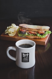 A steaming cup of coffee in a white mug with a branded logo sits in the foreground on a wooden table. Behind the mug, a wooden tray holds a grilled sandwich with lettuce, tomatoes, and cheese. A small pile of potato chips accompanies the sandwich, adding a casual and appetizing touch.