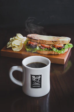 A warm, inviting shot of a turkey club sandwich next to a steaming cup of cappuccino on a wooden table.