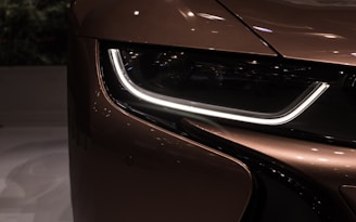 A close-up view of the front headlight of a modern car. The headlight has a sleek design with a prominent LED strip. The car's surface has a glossy, reflective finish, giving it an elegant and futuristic appearance.
