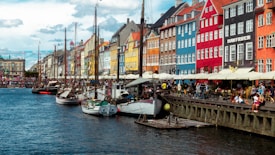 A vibrant waterfront scene featuring a row of colorful, historic buildings along a canal. Numerous sailing ships are moored by the water, and people are sitting and walking along the quay. Outdoor cafes and umbrellas line the dock area, contributing to a lively and bustling atmosphere with blue skies and a few clouds overhead.