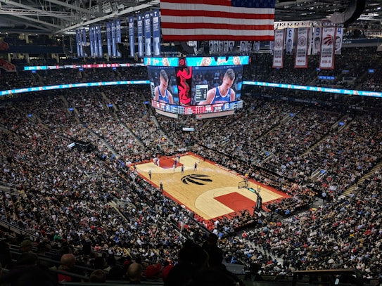 An indoor basketball arena is filled with a large crowd watching a live basketball game. The stadium is adorned with numerous banners and a large American flag. A central scoreboard displays detailed information about the game, including a close-up of a player.