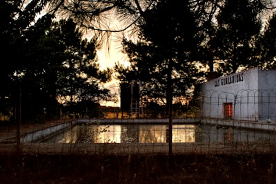 A swimming pool surrounded by a metal fence is seen amidst a rural setting with tall trees framing the area. The trees are silhouetted against the sky, suggesting it is either dusk or dawn. A building with the words 'LAS GUALARIDAS' on it stands nearby, with one window dimly illuminated.
