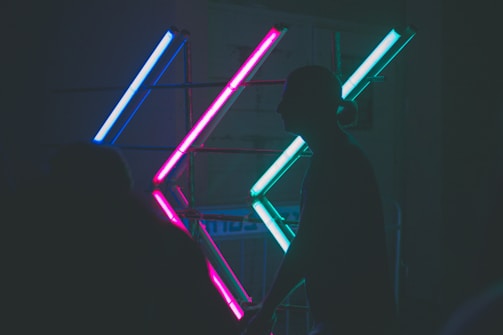 Neon lights arranged in intersecting patterns create a vibrant display against a dark background. The silhouette of a person is visible in the foreground, adding depth to the scene.