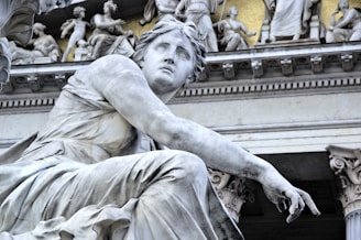 A classical marble statue of a seated figure, looking contemplative, surrounded by intricate architectural details and other sculpted figures in the background.