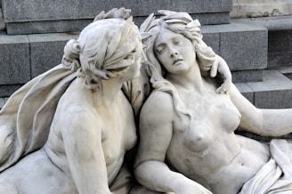 Two intricately carved marble statues depict a tender and intimate scene. The statues are positioned closely together on a stone step structure. One figure has their arm around the other, who appears to be reclining with a serene expression.