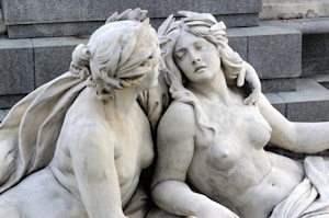 Two intricately carved marble statues depict a tender and intimate scene. The statues are positioned closely together on a stone step structure. One figure has their arm around the other, who appears to be reclining with a serene expression.