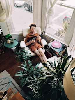 A person is relaxing in a comfortable chair while holding a small string instrument, possibly a ukulele. Surrounding them are various houseplants adding a lush, green ambiance to the room. A round table beside the chair holds a green lamp and a small decorative item. A turntable with a pink vinyl record is on the floor. The room has large windows with curtains that allow natural light to flood in, and a street with parked cars is visible outside.