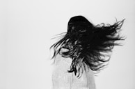 A person with long, dark hair is captured in motion, with hair dramatically spread out to the side against a plain white background. The individual is wearing a light-colored knit sweater and is turned slightly away from the camera, creating a dynamic sense of movement.