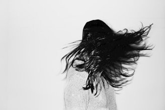 A person with long, dark hair is captured in motion, with hair dramatically spread out to the side against a plain white background. The individual is wearing a light-colored knit sweater and is turned slightly away from the camera, creating a dynamic sense of movement.