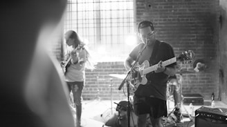 A black and white image of a small band performing in a dimly lit venue with brick walls. Two guitarists are playing, one in the foreground singing into a microphone while playing an electric guitar, and another in the background focused on his instrument. A drum set is visible behind them with a bright light adding dramatic shadows and highlights to the scene.