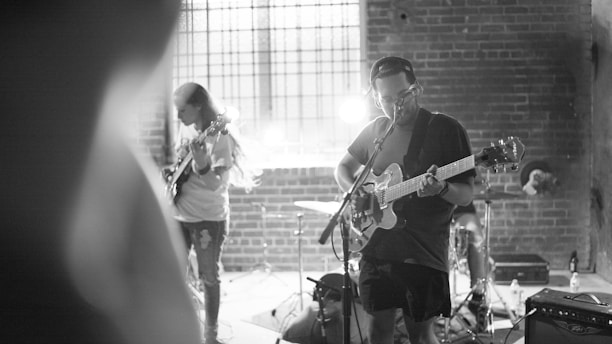 A black and white image of a small band performing in a dimly lit venue with brick walls. Two guitarists are playing, one in the foreground singing into a microphone while playing an electric guitar, and another in the background focused on his instrument. A drum set is visible behind them with a bright light adding dramatic shadows and highlights to the scene.