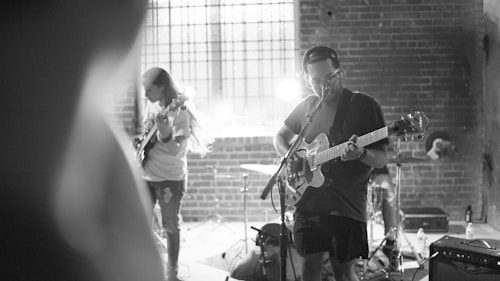 A black and white image of a small band performing in a dimly lit venue with brick walls. Two guitarists are playing, one in the foreground singing into a microphone while playing an electric guitar, and another in the background focused on his instrument. A drum set is visible behind them with a bright light adding dramatic shadows and highlights to the scene.