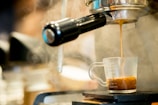 A steaming shot of espresso pours from an espresso machine into a clear glass measuring cup on a drip tray. The rich brown color of the coffee contrasts with the shiny metallic surfaces of the machine.