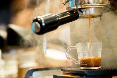 A steaming shot of espresso pours from an espresso machine into a clear glass measuring cup on a drip tray. The rich brown color of the coffee contrasts with the shiny metallic surfaces of the machine.
