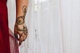 An intricately designed henna pattern is applied to a hand, featuring floral and circular motifs. The hand is positioned against a backdrop of sheer white fabric with hanging decorative strands. A red fabric drapes over the wrist, adding a touch of color.