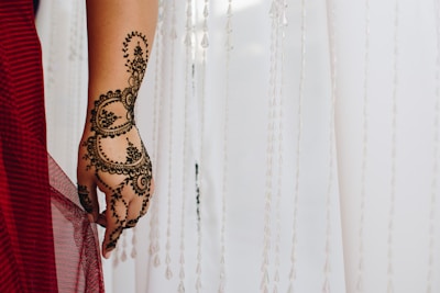 An intricately designed henna pattern is applied to a hand, featuring floral and circular motifs. The hand is positioned against a backdrop of sheer white fabric with hanging decorative strands. A red fabric drapes over the wrist, adding a touch of color.