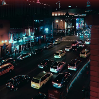 A lively urban street at night with heavy traffic. Bright neon signs illuminate the buildings, creating a vibrant atmosphere. People are walking along the sidewalk, and numerous cars are moving on the road, suggesting a bustling nightlife scene. The architecture features a mix of modern and older buildings, contributing to the city’s dynamic character.