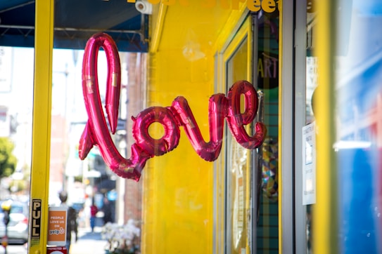 Pink balloon letters spell out the word 'love' against a bright yellow wall. The reflection of these letters can be seen in the glass window of a storefront. The scene is set on a sunny street with a blurred background of pedestrians and urban architecture.