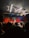 A live music performance in a dimly lit venue, with a vibrant curtain backdrop. A singer is performing on stage, accompanied by musicians. The audience, some recording with their phones, watches closely from the foreground. Stage lights create a dynamic and colorful atmosphere.