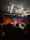 A live music performance in a dimly lit venue, with a vibrant curtain backdrop. A singer is performing on stage, accompanied by musicians. The audience, some recording with their phones, watches closely from the foreground. Stage lights create a dynamic and colorful atmosphere.