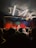 A live music performance in a dimly lit venue, with a vibrant curtain backdrop. A singer is performing on stage, accompanied by musicians. The audience, some recording with their phones, watches closely from the foreground. Stage lights create a dynamic and colorful atmosphere.