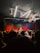 A live music performance in a dimly lit venue, with a vibrant curtain backdrop. A singer is performing on stage, accompanied by musicians. The audience, some recording with their phones, watches closely from the foreground. Stage lights create a dynamic and colorful atmosphere.