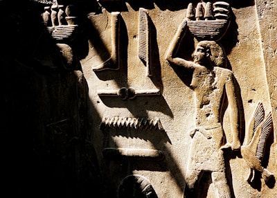 An ancient Egyptian carving depicts figures and hieroglyphs on a stone surface. The artwork features two human figures, each carrying baskets on their heads. The surrounding symbols and objects include a bird and various hieroglyphic elements, all intricately carved with attention to detail.