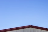 A simple structure with a metal roof against a clear blue sky. The roof has a maroon-colored edge, and the walls beneath appear to be light beige or cream-colored.