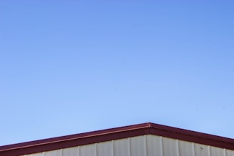 A simple structure with a metal roof against a clear blue sky. The roof has a maroon-colored edge, and the walls beneath appear to be light beige or cream-colored.