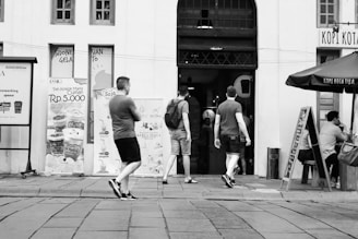 Three men are walking towards an entrance of a building with various promotional posters on the wall. One of them is carrying a backpack. There is a seating area with a man seated at a table under an umbrella labeled 'Kopi Kota Tua'. The setting seems to be urban, with a paved walkway in front.