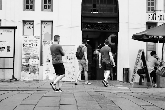 Three men are walking towards an entrance of a building with various promotional posters on the wall. One of them is carrying a backpack. There is a seating area with a man seated at a table under an umbrella labeled 'Kopi Kota Tua'. The setting seems to be urban, with a paved walkway in front.