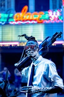 A person with intricate body paint and a futuristic weapon slung over their shoulder stands in front of a brightly lit neon sign. The individual wears a white shirt, black tie, and sunglasses, exhibiting a cyberpunk aesthetic.