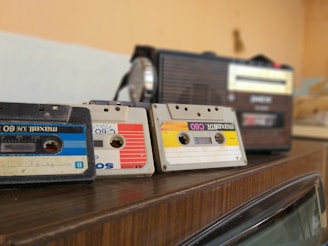 Three vintage audio cassette tapes in various colors are placed on a wooden surface next to a retro-style radio. The cassettes and radio create a nostalgic ambience.
