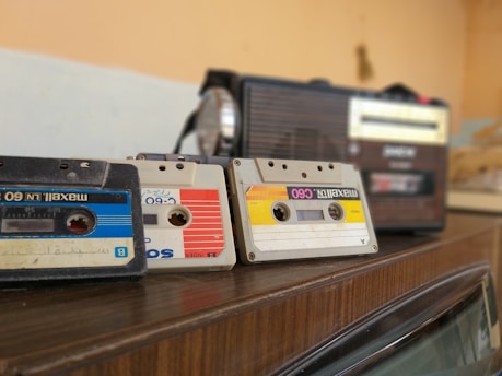 Three vintage audio cassette tapes in various colors are placed on a wooden surface next to a retro-style radio. The cassettes and radio create a nostalgic ambience.
