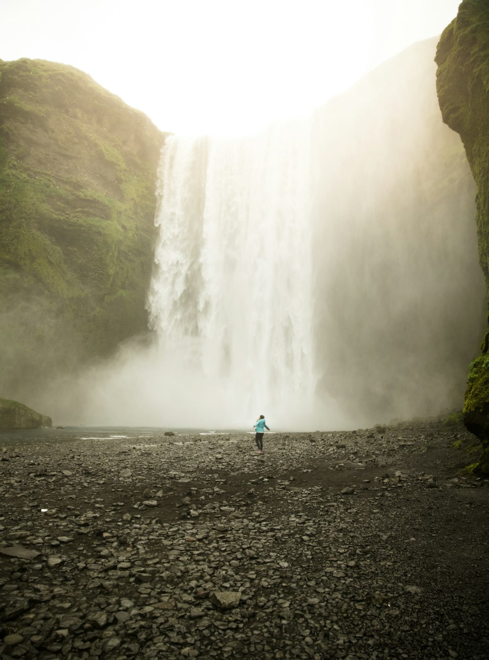 person in blue jacket standing on gray rocky ground near waterfalls during daytime