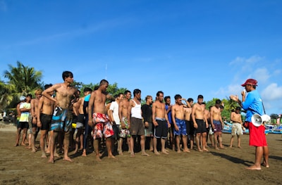 A group of men are standing together on a sunny beach, listening to a man who appears to be instructing them. The instructor is wearing a cap and holding a megaphone. The men are casually dressed in shorts and are mostly bare-chested, suggesting the setting is informal and possibly related to a beach activity or sport. Palm trees and a clear blue sky provide the backdrop.
